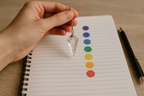 Hand holding a crystal pendulum over a notebook with color swatches on a wooden surface.